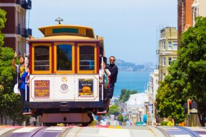 San Francisco, United States - May 19, 2016: Approaching iconic cable car is full of outside hanging and platform standing tourists with bay water background in sunny Powell Street in California. Horizontal